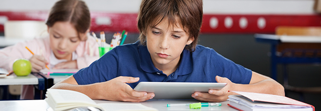 Elementary schoolboy using digital tablet with classmate studyin